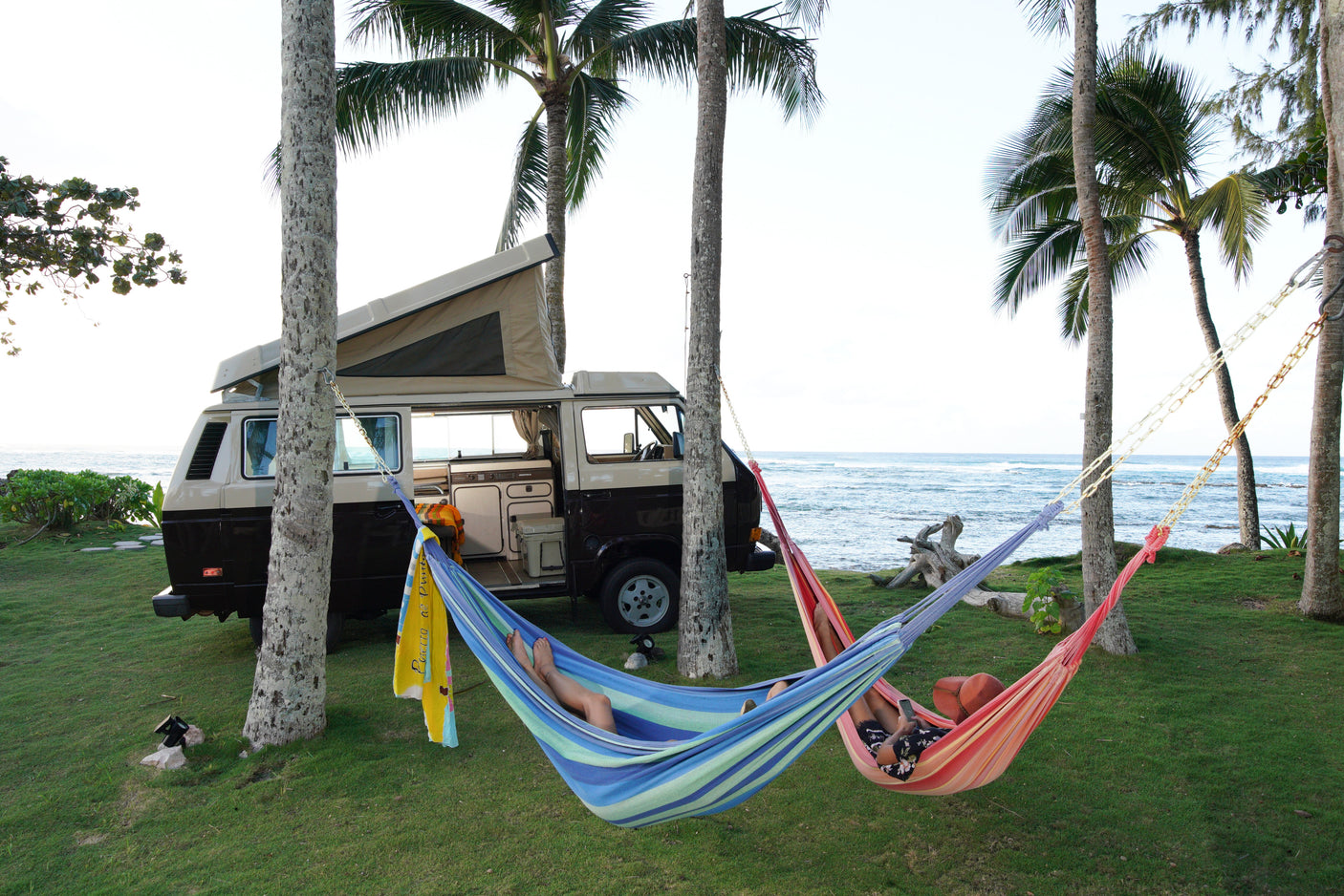 Your RV rental adventure: A camper van by the ocean, two people in hammocks under palms. Perfect beach relaxation!