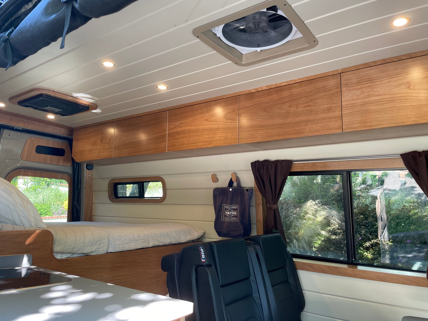 Cozy RV rental interior with wood cabinets, a comfy bed, black seats, ceiling fan, and natural light from windows.