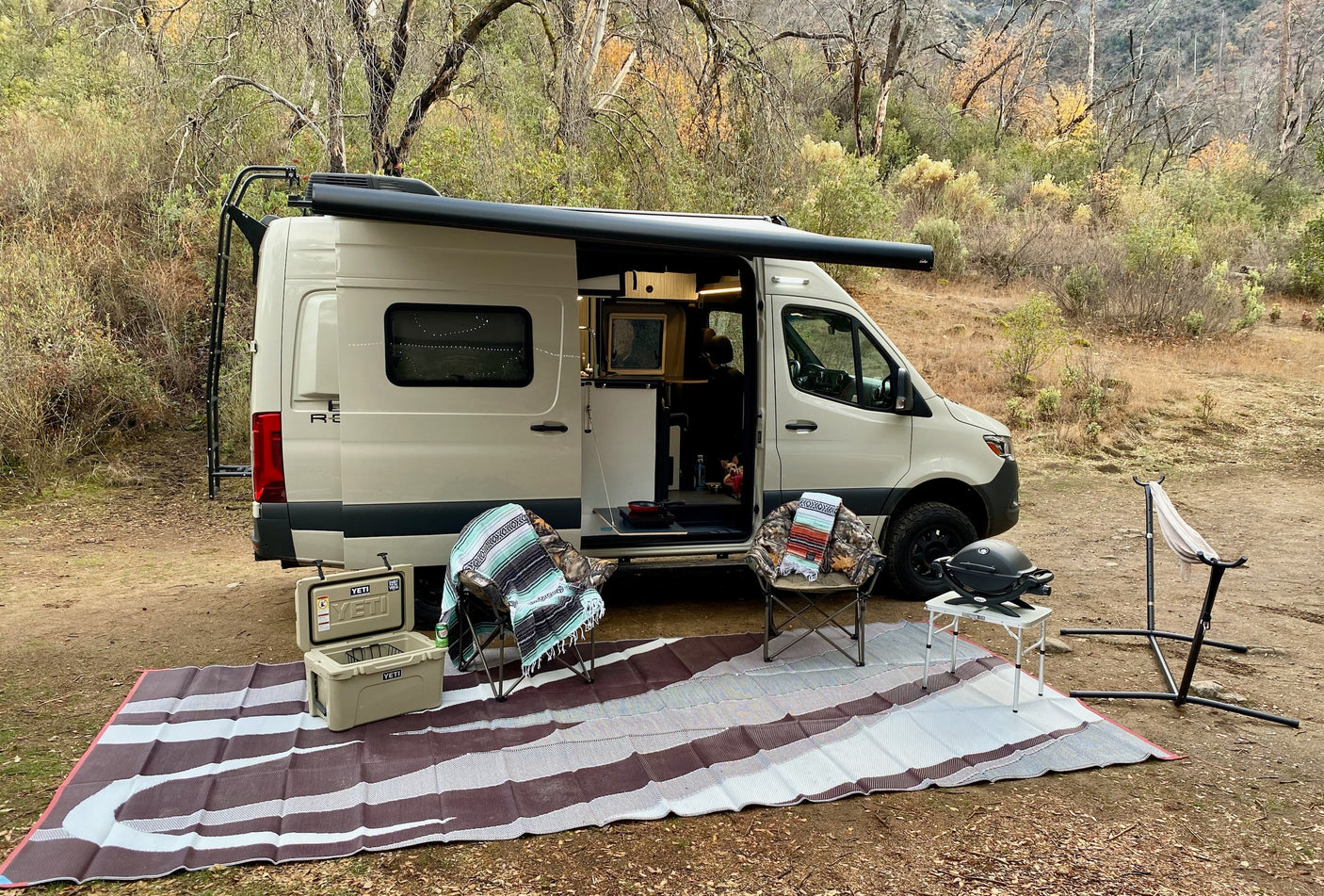 Tan rv rental camper van with awning, set up for outdoor camping. Features chairs, Yeti cooler, and grill in a natural setting.