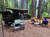 Overlanding setup: dark camper trailer & Jeep. People by a campfire in a forest. Ideal RV rental for outdoor living!