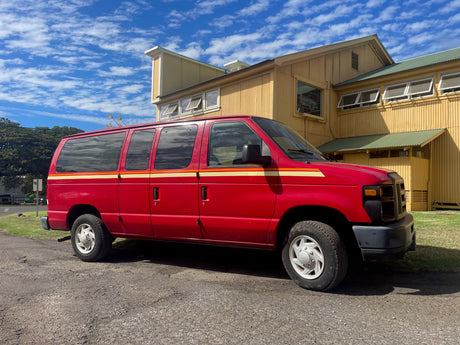 A red van, reminiscent of an RV rental, parked alongside a yellow building under blue, cloudy skies.