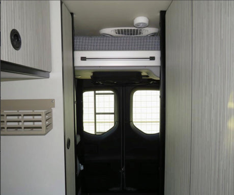 Compact RV rental interior: overhead bunk, fan, light, storage cabinetry, and rear doors with grid windows.