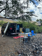 Explore with an RV rental! Camper van with surfboard, portable stove, and chairs set up on a rocky shore under a large tree.