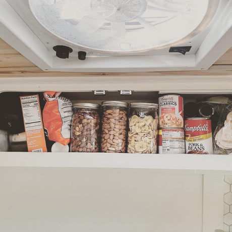 Inside an rv rental, an overhead pantry is well-stocked with canned goods, jars of snacks, and chips, ready for a road trip.