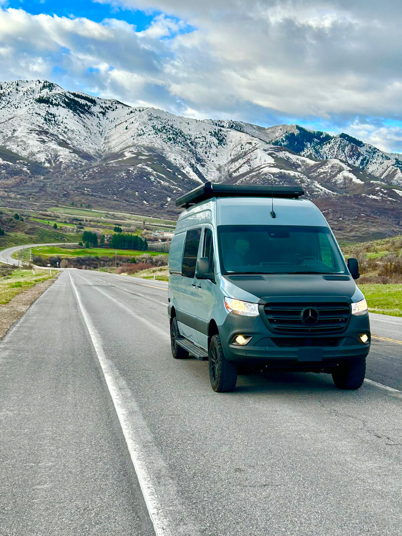 Adventure awaits! This sleek **RV rental** camper van on a scenic mountain road with snow-capped peaks under a cloudy sky. — photo 2
