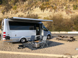 Coastal RV rental setup with Airstream camper van, awning, chairs, cooler & picnic table on sand near cliff.