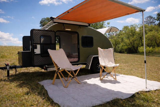Experience nature with this olive green teardrop camper RV rental, featuring an orange awning, open door, and two chairs on a rug outdoors.