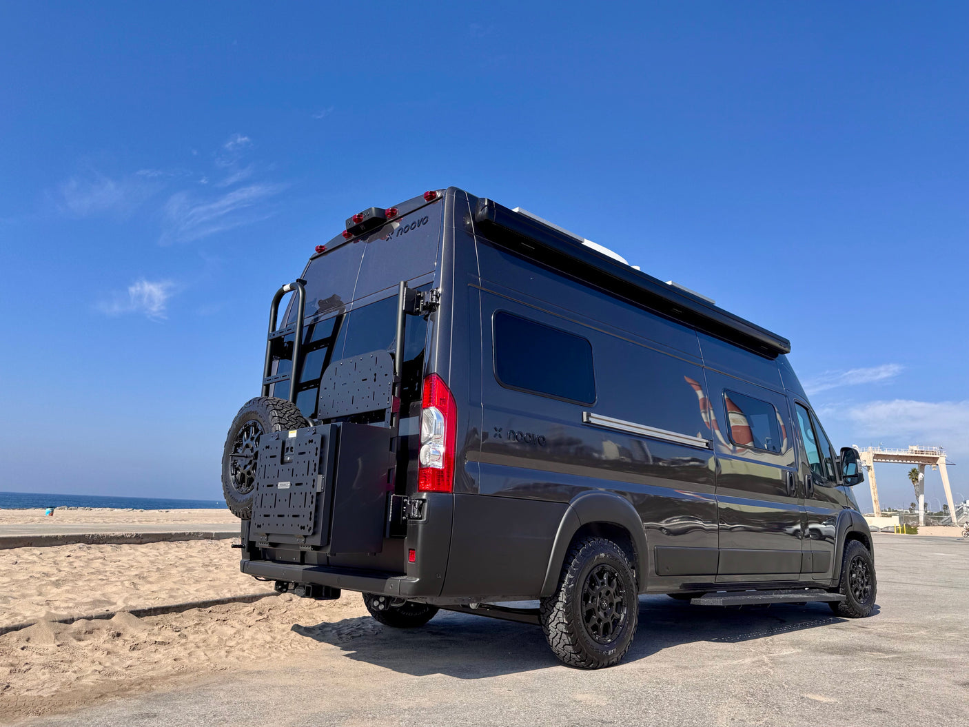 Dark gray 'X noovo' RV rental parked by a sandy beach. Rear view shows ladder, spare tire, and modular storage.