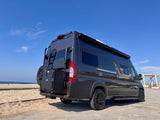 Dark gray 'X noovo' RV rental parked by a sandy beach. Rear view shows ladder, spare tire, and modular storage.