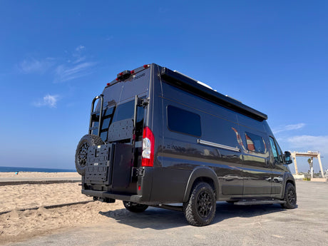 Dark gray 'X noovo' RV rental parked by a sandy beach. Rear view shows ladder, spare tire, and modular storage.