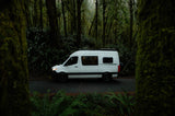 A white RV rental camper van parked on a forest road amidst moss-laden trees and vibrant ferns.