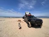 Black modified pickup truck with rooftop tent & solar, dogs, and ocean views on a sandy beach. Great for an RV rental adventure!