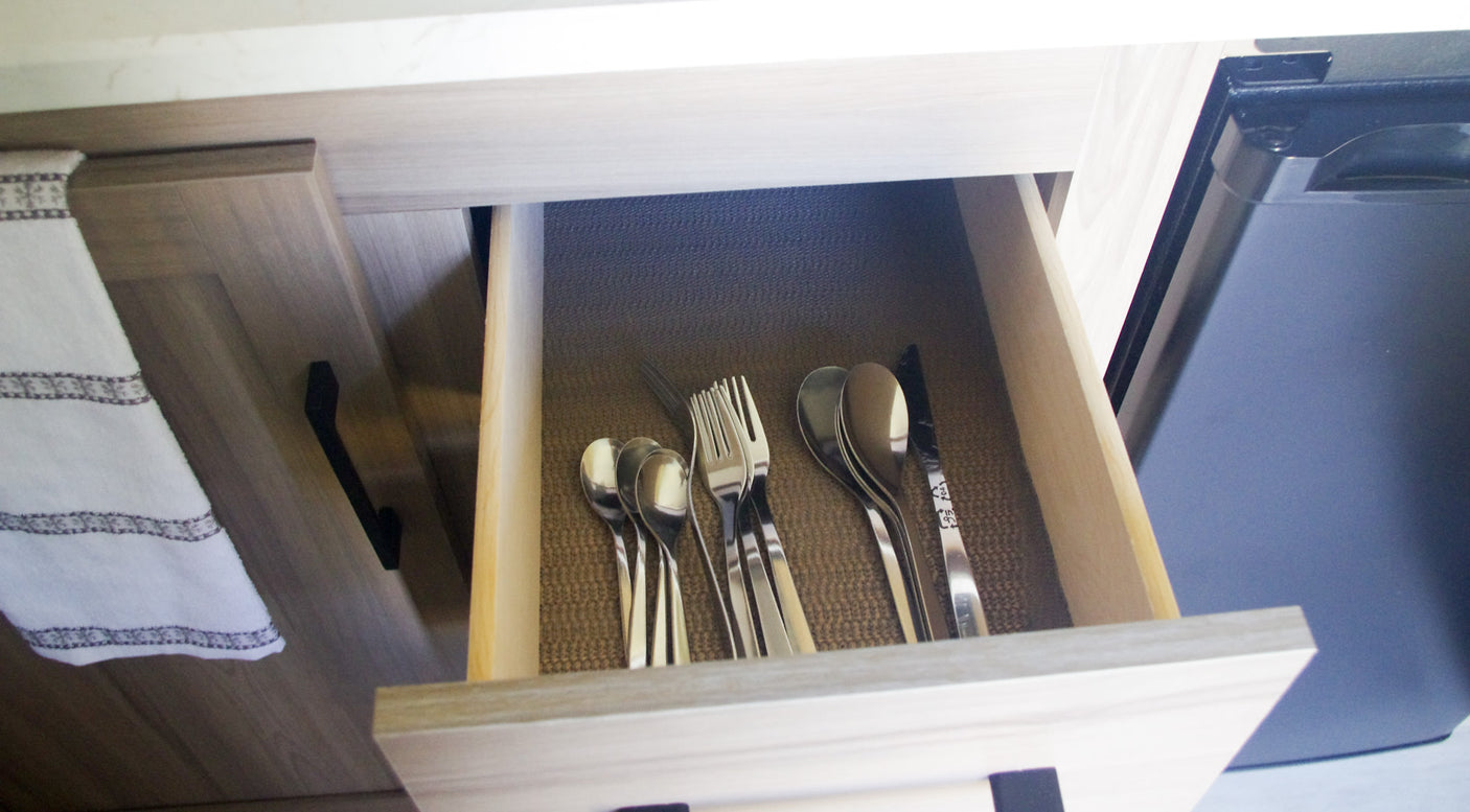Inside an RV rental, an open kitchen drawer reveals neatly organized silverware, next to a hanging towel and fridge.