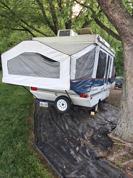 White pop-up RV rental, extended for camping, with a black ground cover & green trees.