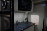 Modern RV rental interior shows a compact kitchenette: black speckled counter, sink, microwave, and window blinds.