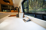 Inside a modern rv rental: kitchen counter with coffee, pop-up power, and serene forest views from the bed area.