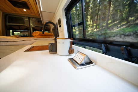 Inside a modern rv rental: kitchen counter with coffee, pop-up power, and serene forest views from the bed area.