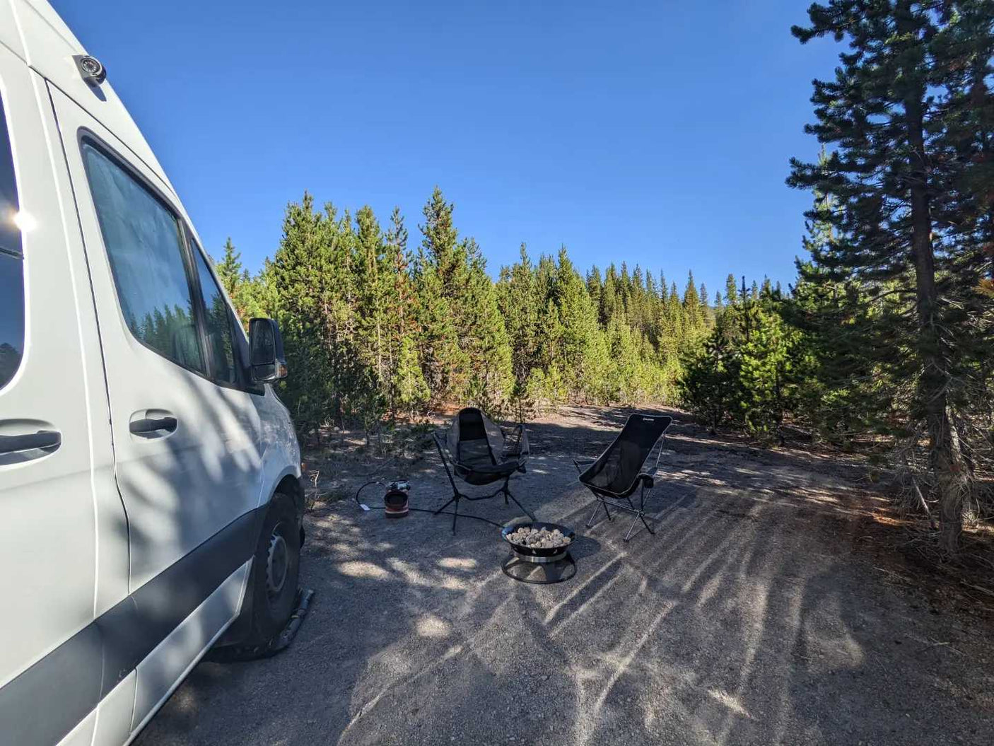 RV rental camping in a serene forest. White campervan, two chairs, and fire pit are set up for a relaxing outdoor experience.