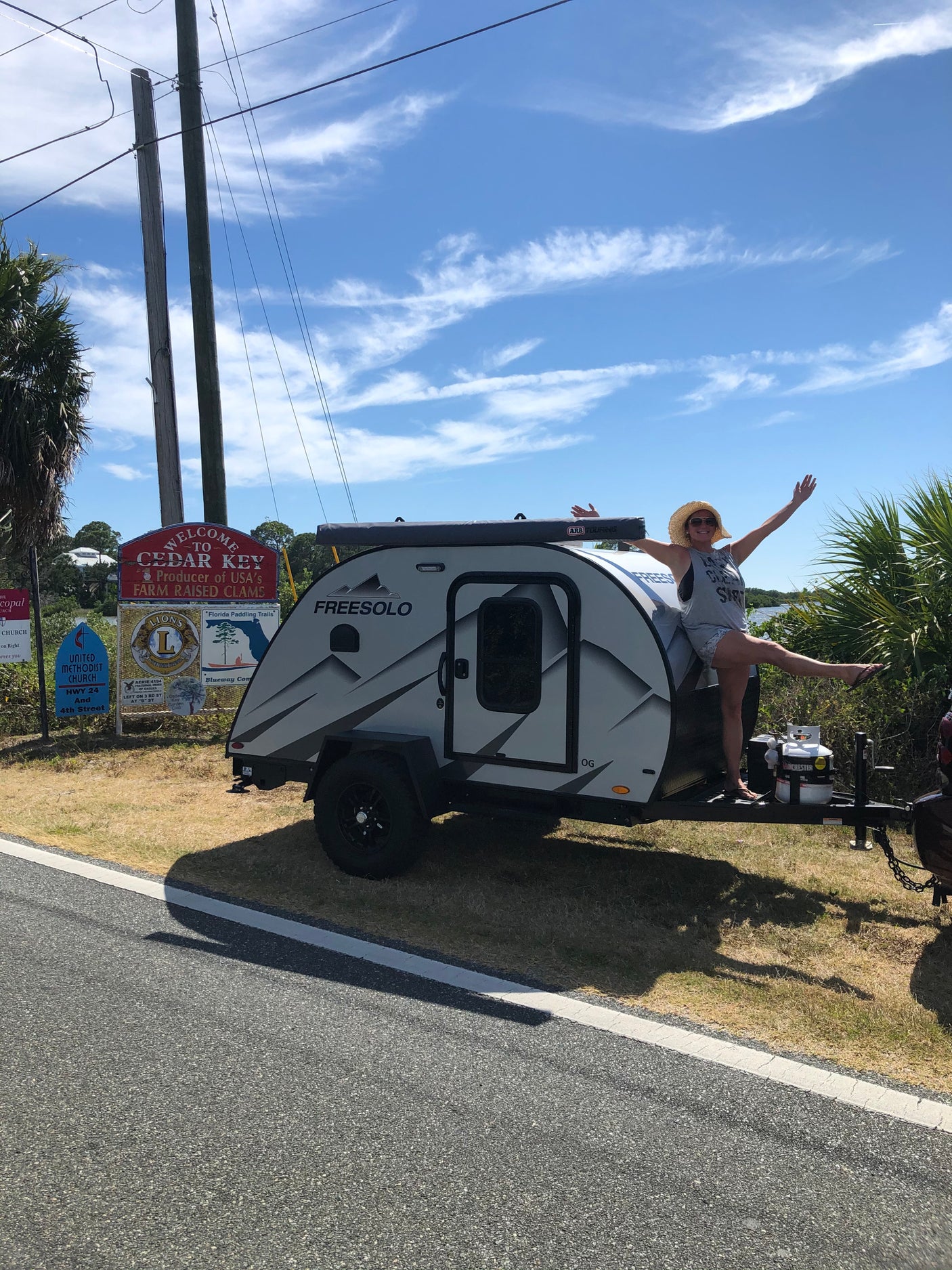 A woman poses on a Freesolo teardrop travel trailer near a "Welcome to Cedar Key" sign. Ideal for an RV rental trip!