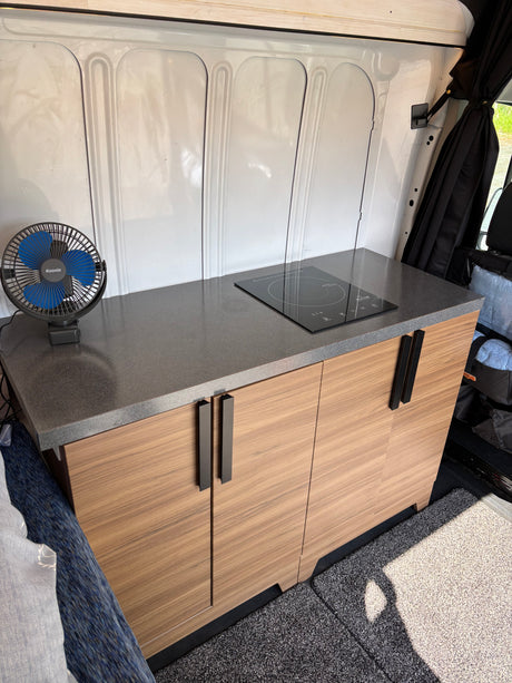 Compact RV rental kitchenette: sleek counter, induction cooktop, and modern wood cabinets in a camper van interior.