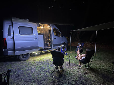 RV rental camper van brightly lit at night. Friends relax in chairs under the awning, enjoying a cozy outdoor camp evening.