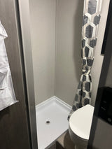 Inside a compact RV rental bathroom: shower stall with white pan, drain, and gray walls, plus a toilet and patterned shower curtain.