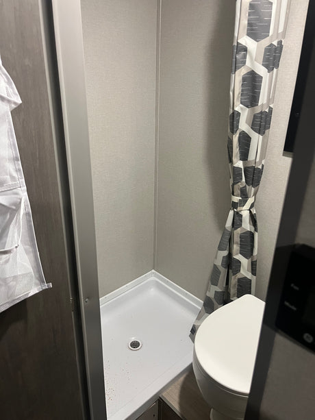 Inside a compact RV rental bathroom: shower stall with white pan, drain, and gray walls, plus a toilet and patterned shower curtain.
