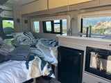 Interior of an RV rental featuring two beds with patterned bedding, a compact kitchen, and scenic mountain views outside.