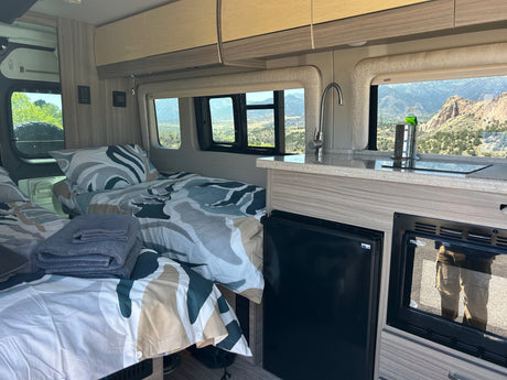 Interior of an RV rental featuring two beds with patterned bedding, a compact kitchen, and scenic mountain views outside.