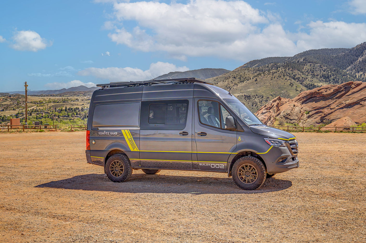 This rugged adventure van RV rental, perfect for off-road travel, is parked in a dusty mountain landscape under a blue sky.