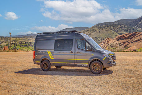 This rugged adventure van RV rental, perfect for off-road travel, is parked in a dusty mountain landscape under a blue sky.