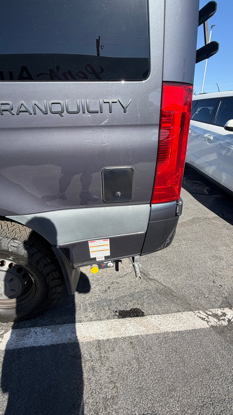 Close-up of a gray Tranquility RV rental's rear, revealing a taillight, utility port, tire, and propane connections.