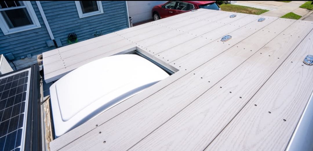 Overhead view of an RV rental roof, showcasing a light-colored wooden deck, a white skylight, and solar panels for off-grid travel.