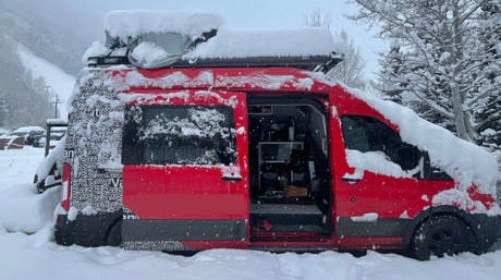 A red camper van, an RV rental, covered in snow in a mountain setting. Its side door is open, revealing the interior living space.