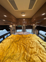 Cozy rv rental bedroom interior featuring a comfortable yellow bed, overhead storage, and windows, perfect for travel.