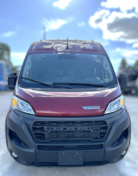 Front view of a red Winnebago RV rental van, based on a Ram ProMaster chassis, parked outdoors under a blue sky.