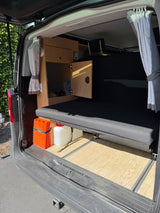 RV rental: Rear interior of a camper van, featuring a bed, wood cabinets, curtains, and stored gear like traction boards.