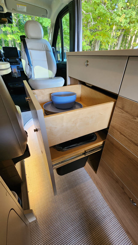 Enjoy your RV rental! Interior view of a passenger seat, pull-out kitchen drawers with dishes, and a waste bin.