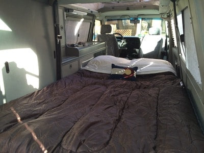 Spacious RV rental bed setup with brown comforter & Colorado pillow. Shows built-in storage/kitchen area. — photo 13