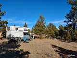 XP Camper truck camper parked among pine trees. Ready for your next outdoor RV rental adventure, with chairs set up.