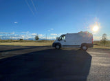A white cargo van, perfect as an RV rental, parked on asphalt. Bright sun, distant mountains, & open fields under a blue sky.