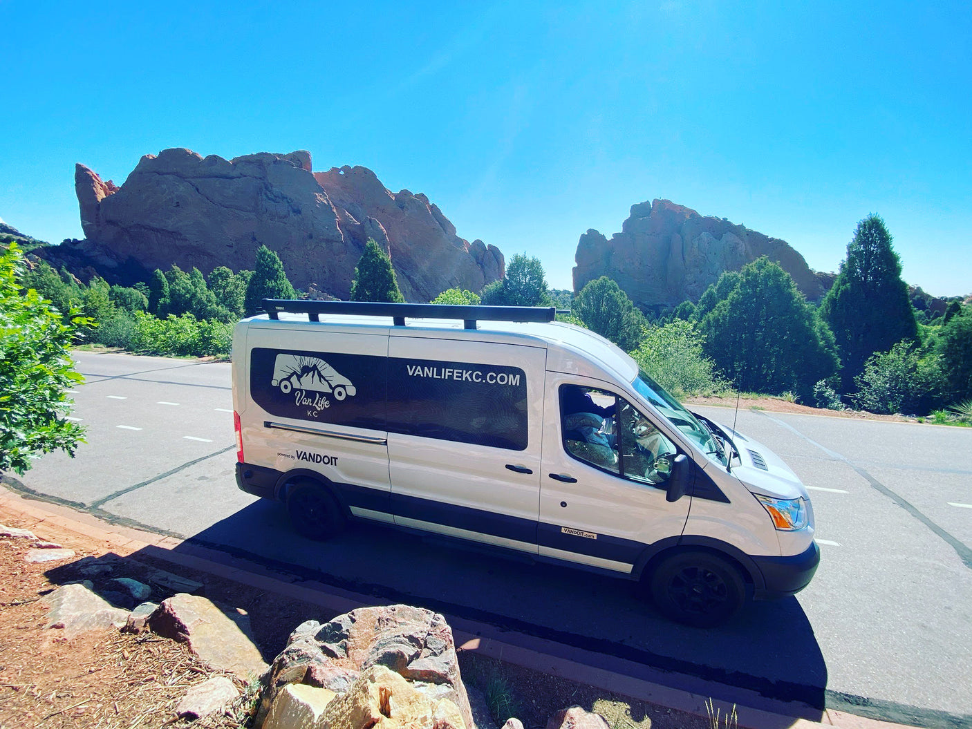 White RV rental van parked near majestic red rock formations & trees under a bright blue sky. Explore nature in comfort.