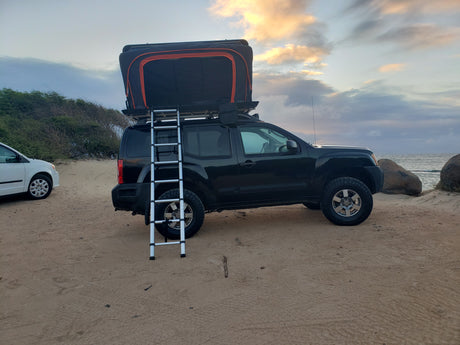 This black SUV with a rooftop tent and ladder, parked on a sandy beach, is perfect for an RV rental outdoor adventure.