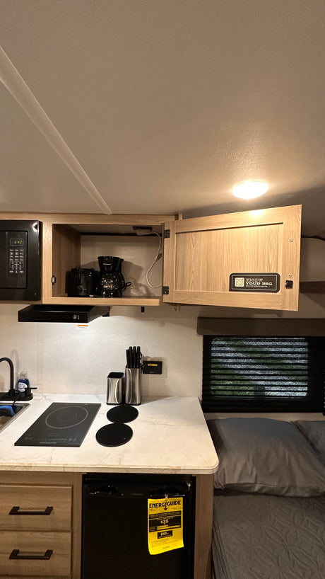 This RV rental's compact kitchen features a microwave, coffee maker, induction cooktop, and mini-fridge, next to a bed.