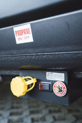 Close-up of an RV rental propane system: "ON/OFF" switch, yellow connection cap, and liquid fill valve with safety warnings.