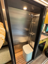 Compact RV rental bathroom: sleek stainless steel shower with wood slats, portable toilet, and integrated living/kitchen area.