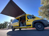 Yellow camper van with extended awning, side door open, under a blue sky. This RV rental is ready for your travel adventures.