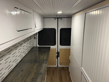 Bright RV rental conversion van interior with white ceiling, custom cabinets, patterned walls, and wood flooring.
