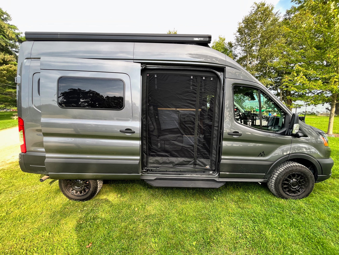A sleek grey conversion van, set up as an RV rental, parked on grass with its side screen door open for fresh air.