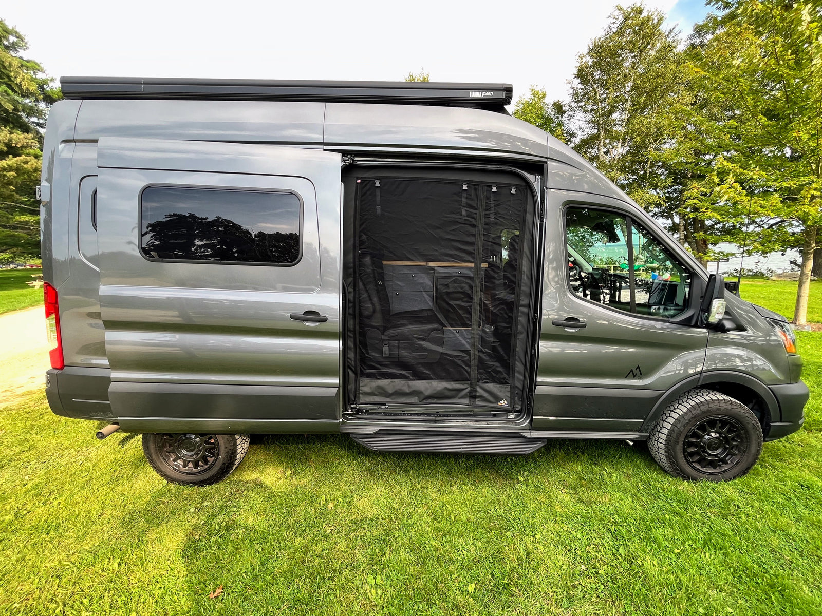 A sleek grey conversion van, set up as an RV rental, parked on grass with its side screen door open for fresh air.